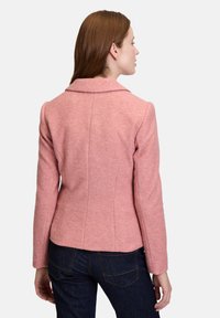 Vaaleanpunainen villasekoitteinen takki, jossa on kaulus, istuva malli ja pitkät hihat. Näkyvät takasaumat ja sileä pinta. Pukeutunut tummansinisiin farkkuihin.