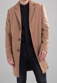 Homme portant un manteau camel à boutons sur une chemise noire à boutons et un jean foncé, devant un fond gris uni.