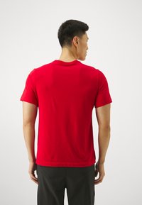 Camiseta vermelha de mangas curtas com decote redondo, feita de tecido suave. Bainha costurada na cintura, usada com calções pretos, vista de costas.