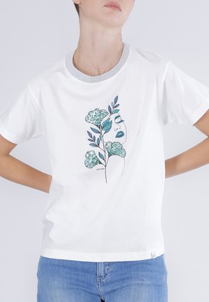 Henkilö, jolla on valkoinen t-paita, jossa on hopeareunainen kaulus ja edessä vihreä ja sininen abstrakti kasvojen ja kukkakuvion yhdistelmä.