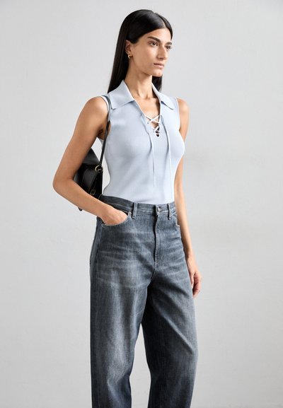 Lys blå ribbet ermeløs topp med krage og snøring, kombinert med høyt liv mørkegrå denimjeans. Modellen holder en svart veske.