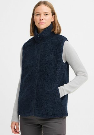 Gilet en polaire bleu marine avec col montant, fermeture éclair à l'avant et poches latérales. Tissu texturé et doux avec une apparence douillette. Superposé sur un pull léger.