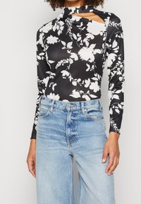 Zwarte långmouwtop met wit bloemmotief, voorzien van een opstaande kraag en uitsnijding. Gecombineerd met lichtblauwe high-waisted jeans.