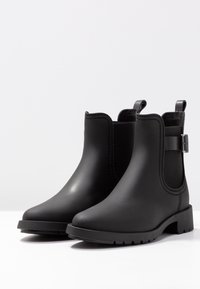 Bottes Chelsea noires en caoutchouc avec des panneaux élastiques sur les côtés, hauteur cheville, bout arrondi, semelle texturée et languettes arrière pour un enfilage facile.