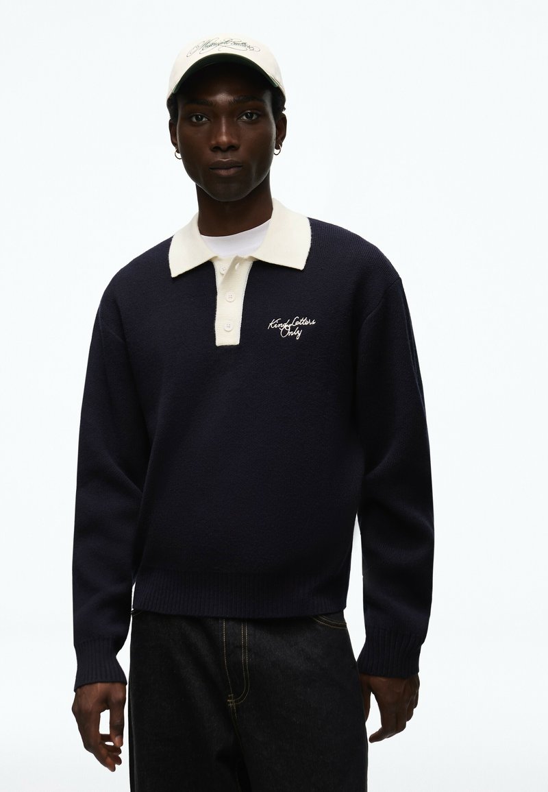 Jeune homme portant un pull bleu marine avec un col crème et un texte brodé, jean foncé, casquette blanche et petites boucles d’oreilles créoles sur fond blanc.