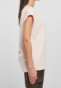 Kobieta przedstawiona z boku, ubrana w jasnoróżowy bezrękawnik i ciemnozielone spodnie cargo na jednolitym szarym tle.