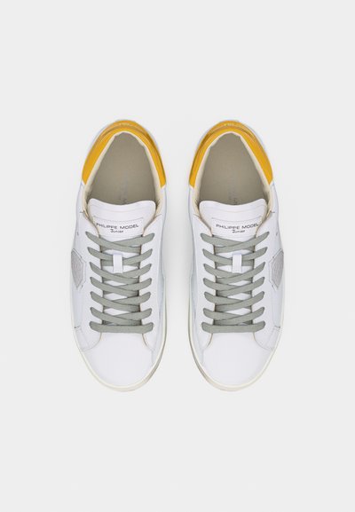 Paire de baskets basses blanches avec des lacets gris et des accents jaunes au talon, étiquetées "Philippe Model Junior" sur la languette.
