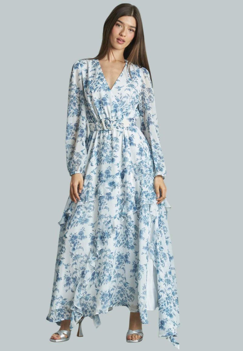 Weißes Maxikleid mit blauen Mustern, langen Ärmeln, V-Ausschnitt, tailliertem Schnitt mit einem Gürtel, geschichteten Rüschendetails und silbernen Sandalen.