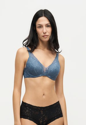 Femme aux longs cheveux foncés portant un soutien-gorge en dentelle bleue et une culotte en dentelle noire, debout devant un fond clair uni.