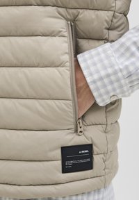 Mano en el bolsillo de una chaqueta beige acolchada con cremallera, usando una camisa a cuadros en tonos azul claro y blanco, con la manga visible.