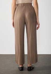 Bruine geplooide broek met wijde pijpen van een gestructureerde stof, elastische tailleband en een vloeiend ontwerp. De zoom valt op de enkel.