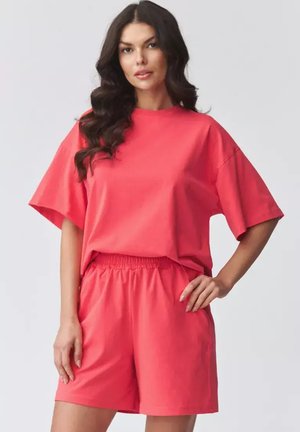 Kobieta o długich ciemnych włosach, ubrana w luźny koralowy t-shirt i dopasowane szorty z elastycznym pasem, stoi z jedną ręką na biodrze.