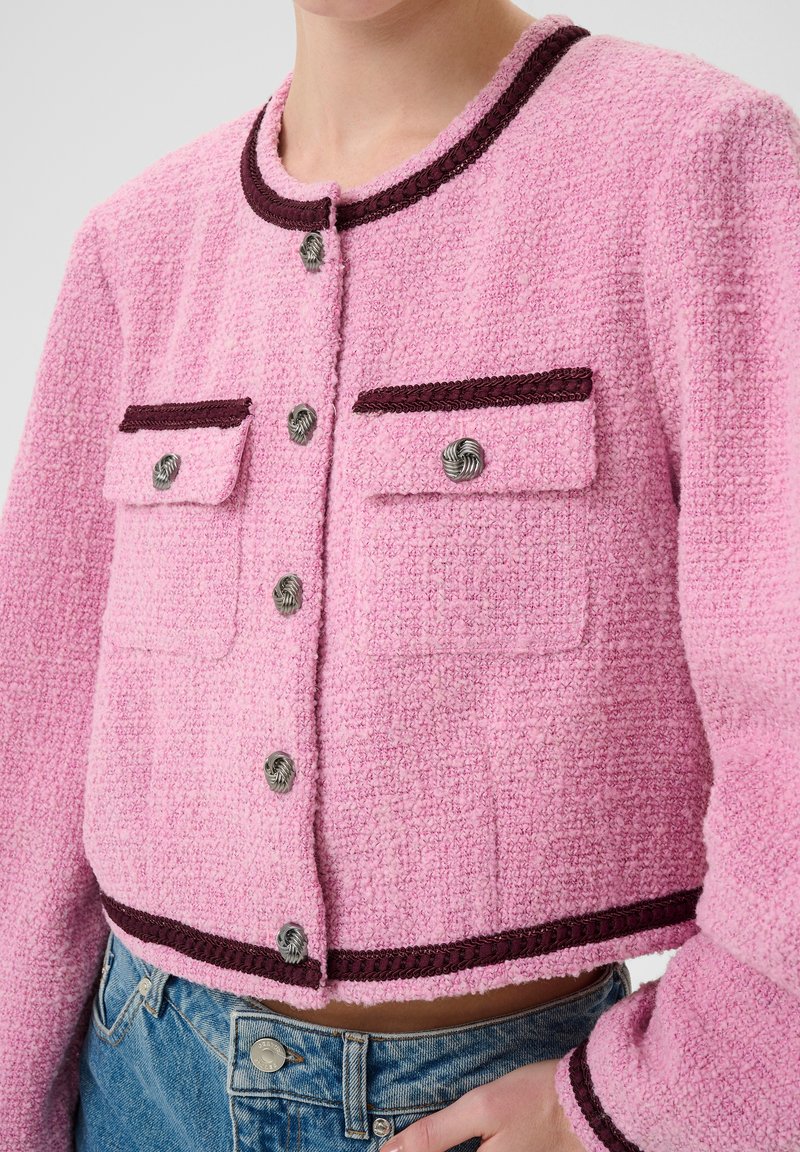 Kort rosa cropped jacka i texturerad tyg, med kontrasterande mörka detaljer, två framfickor med knappar och rund halsringning.