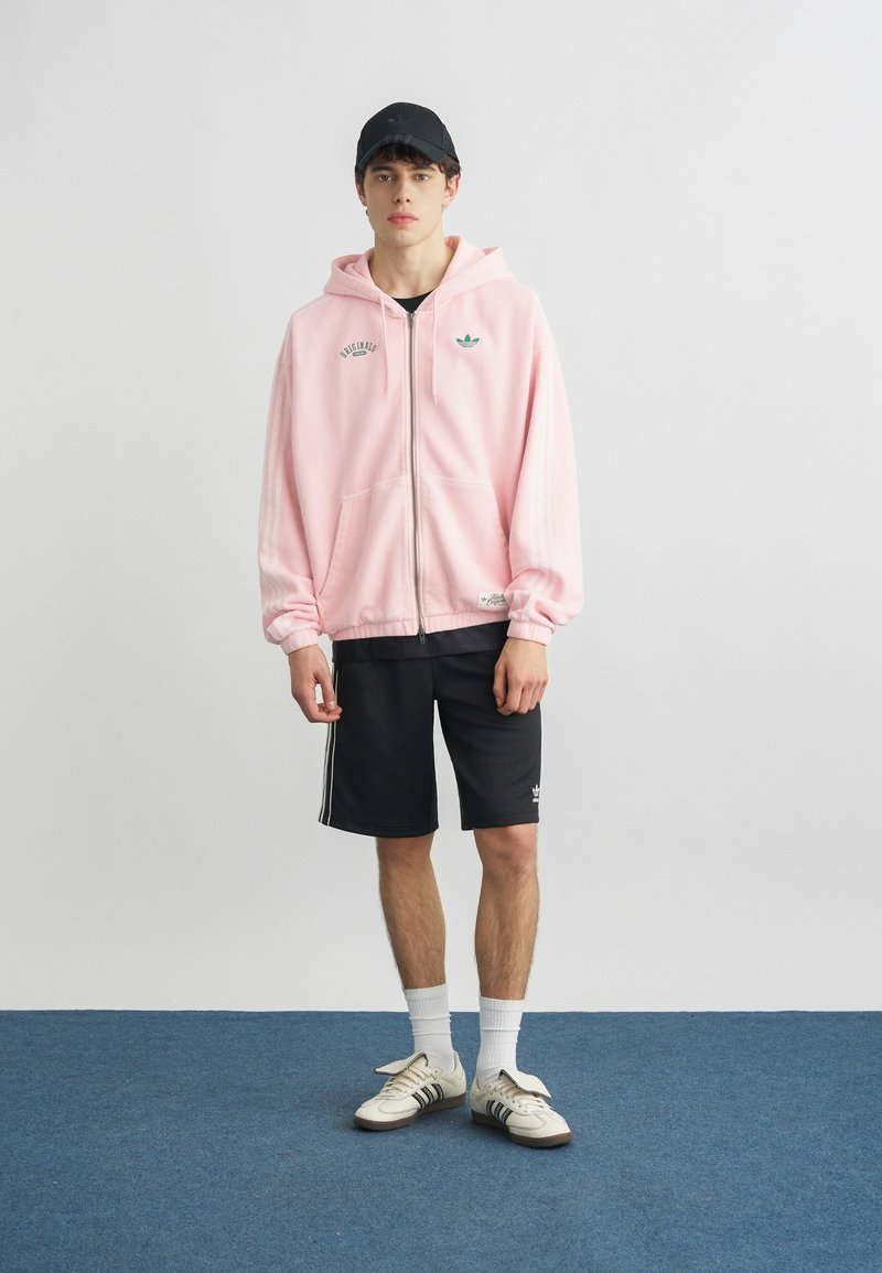 Jeune homme portant un sweat à capuche zippé rose, un short noir, des chaussettes blanches, des baskets blanches et une casquette noire, debout sur un tapis bleu.