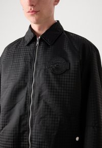 Veste à carreaux noirs avec fermeture éclair, col, et un patch rond texturé sur la poitrine, présentant de subtils motifs géométriques.