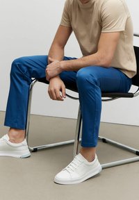 Beige přiléhavé tričko, modré slim-fit kalhoty a bílé tenisky. Kalhoty mají hladkou texturu, zatímco boty mají šněrovací design a gumové podrážky.