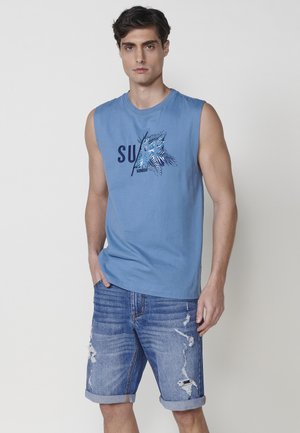 Camicetta blu in cotone senza maniche con grafica "SURF" e design di fronde di palma; abbinata a pantaloni di jeans strappati con risvolti.