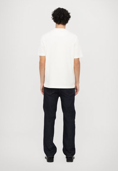 Bela bombažna majica s kratkimi rokavi, v kombinaciji s temno modrimi kavbojkami. Naravnost krojena z diskretnim šivanjem na zadnjih žepih.