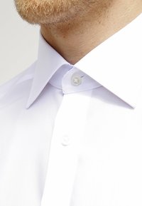 Chemise blanche à col classique, dotée de deux boutons et d'une texture lisse et élégante. Le tissu semble frais et bien ajusté.