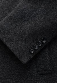 Manica di giacca in lana grigio scuro con motivo a spina di pesce e quattro bottoni neri lungo il polsino. Il tessuto appare morbido e leggermente felpato.