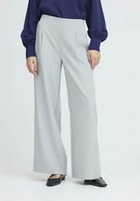 Šviesiai pilki, plačios kojos kelnės su aukštu juosmeniu ir plokščiu priekiu. Audinys atrodo lygus, derinamas su tamsiais, kvadratiniais bateliais.