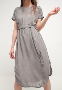 Harmaa mokkanahkamekko lyhyillä hihoilla, koristeellista pitsiä hartioilla, solmittava vyötärö ja pyöristetty helma sivuaukkoineen. Sujuva pinta.