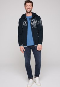 Felpa navy con zip e grafica bianca, t-shirt blu, jeans scuri e sneakers bianche. Presenta polsini a righe e cappuccio con cordino.