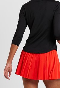 Czarny top z długim rękawem o okrągłym dekolcie, połączony z plisowaną czerwoną spódnicą. Spódnica ma rozkloszowany fason i gładką fakturę.