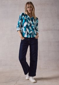 Blusa a maniche lunghe, color teal e blu navy con motivo a V; abbinata a jeans larghi blu scuro e sneakers bianche. Tessuto con texture liscia.