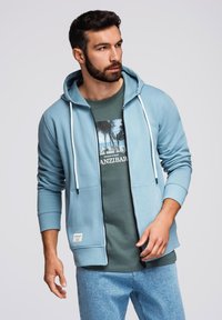 Jasnoniebieska bluza z zamkiem na całej długości i kapturem na sznurku, w połączeniu z zielonym t-shirtem z grafiką palmy, noszona z jasnoniebieskimi dżinsami.