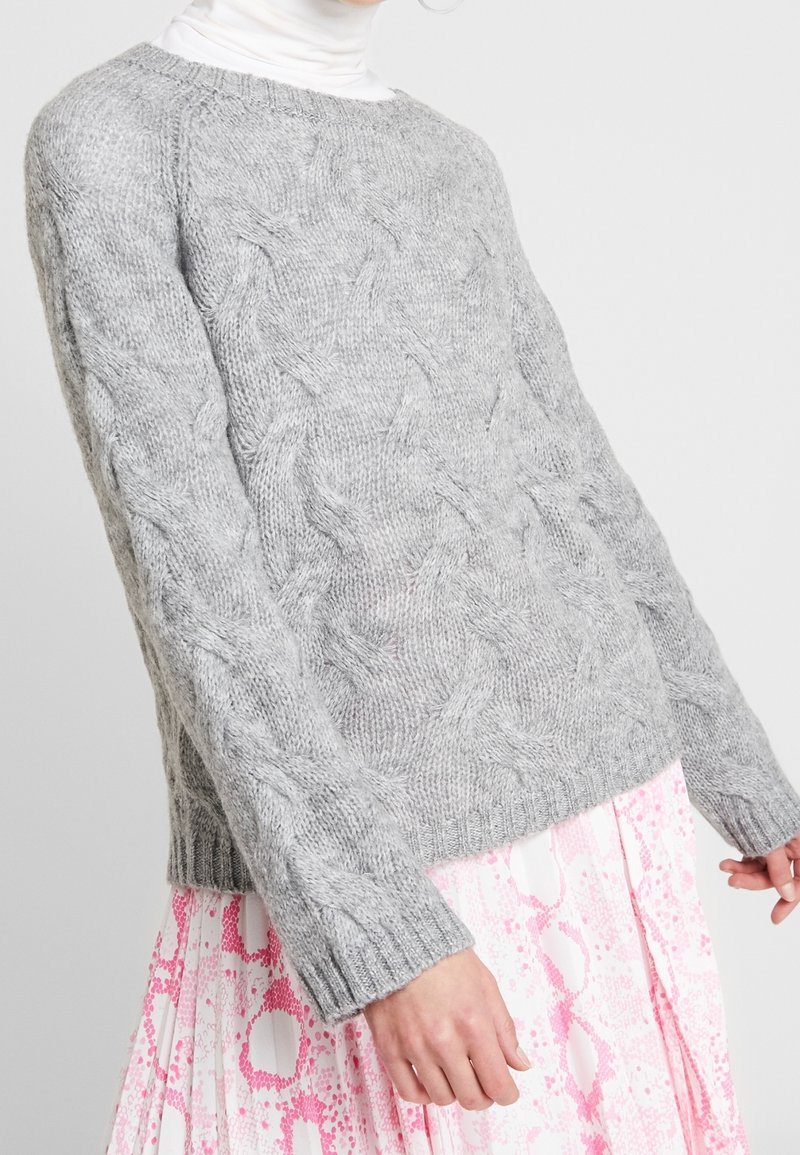 Personne portant un pull gris en maille torsadée sur un col roulé blanc et une jupe plissée à motif rose et blanc.