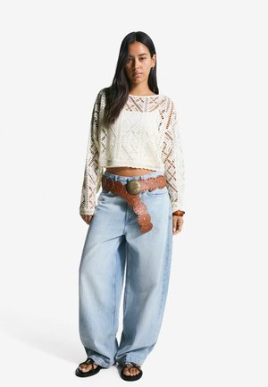 Häkel-Crop-Top in Creme, hoch-taillierte hellblaue Jeans, braiter Ledergürtel mit runden Akzenten und schwarze Sandalen.