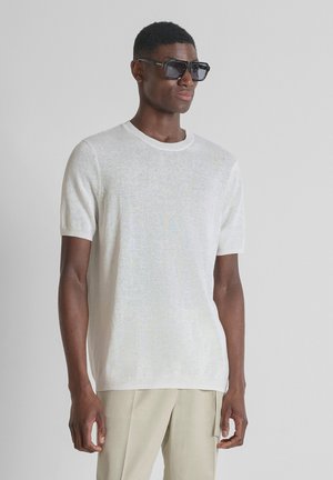 Hombre con gafas de sol, una camisa de punto blanca de manga corta y pantalones beige, de pie frente a un fondo liso de color gris claro.