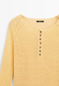 Lysegul strikket langærmet top med rund halsudskæring og syv små brune knapper midt foran.