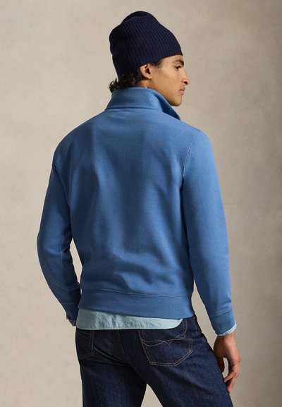 Μπλε βαμβακερό φούτερ με ψηλό γιακά και ριμπ μανσέτες. Φοράται πάνω από ανοιχτό μπλε πουκάμισο, συνδυασμένο με σκούρο τζιν παντελόνι.