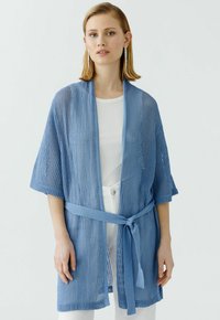 Helles blaues, offen gestricktes Cardigan mit weiten Ärmeln und einem Gürtel in der Taille. Schlichtes Design mit einem strukturierten Muster, getragen über einem weißen Oberteil.