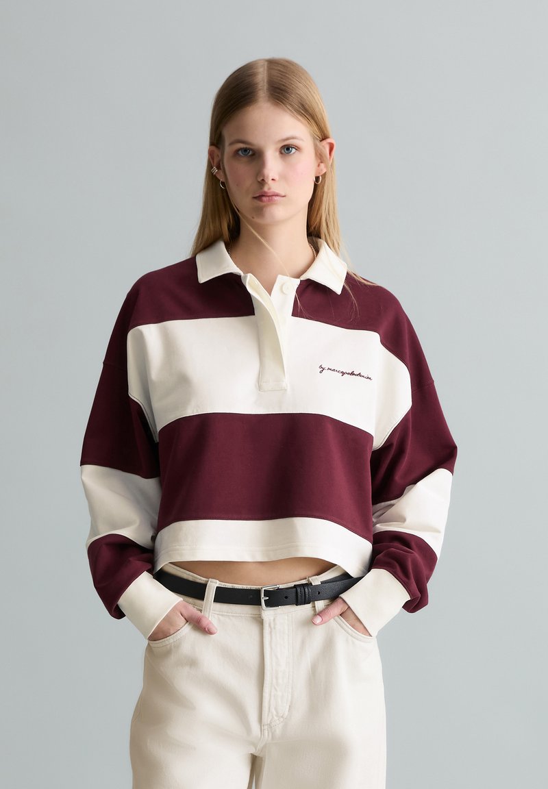 Νέα γυναίκα με μια μωβ και λευκή ριγέ κομμένη φανέλα ράγκμπι, μπεζ παντελόνια και μαύρη ζώνη, στέκεται μπροστά από ένα απλό γκρι φόντο.