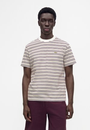 Junger Mann trägt ein weiß-braun gestreiftes kurzärmeliges Hemd mit kleinem grünem Logo und dunkelvioletten Shorts, steht vor einfachem Hintergrund.