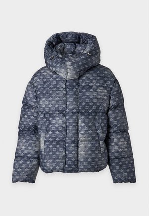Jeune homme portant une doudoune sombre à motifs par-dessus une chemise blanche avec un jean bleu et des baskets blanches, debout devant un fond uni.