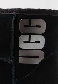 Czarny zamszowy botek z wyraźnym metalowym logo "UGG". Projekt zawiera teksturowane wykończenie i kontrastowe przeszycia wzdłuż szwów.