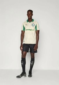 Jogador de futebol alto, em pé, vestindo uma camisola branca e verde, calções pretos, meias altas pretas e sapatilhas pretas, contra um fundo simples.