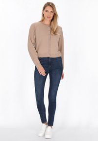 Beige neulecardigan, jossa on pyöreä pääntie ja etupainikkeet, yhdistetty tummansinihin skinny-farkkuihin ja valkoisiin lenkkareihin. Sujuva pinta, lyhyt malli.