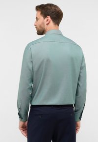 Camicia a maniche lunghe verde chiaro in tessuto testurizzato, con colletto a punta e polsi abbottonati, indossata con pantaloni blu navy.