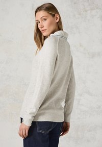Lichtgrijze gebreide trui met een geribbelde textuur en kraag, met een blauwe accent. Model draagt donkere jeans en poseert zijwaarts.