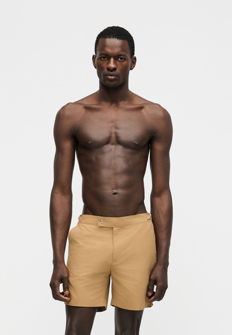 Izmos, sötét bőrű férfi áll előre nézve, homokszínű rövidnadrágot visel, ing nélkül, semleges arckifejezéssel egy egyszerű világos háttér előtt.