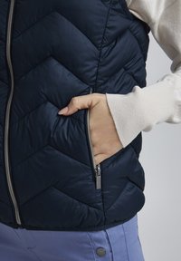 Gilet matelassé bleu marine avec un motif en chevrons, poche zippée et quincaillerie argentée. Manches de pull de couleur claire visibles aux poignets.