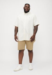 Cămașă bej cu mâneci scurte și nasturi, pantaloni scurți de culoare bej deschis și adidași albi. Cămașa are un logo subtil; pantaloni scurți sunt de lungime până la genunchi și au o croială lejeră.