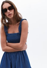 Denim dress with a smocked bodice and ruffled straps, in a medium blue shade. Model wears dark sunglasses and has wavy brown hair.