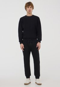 Felpa nera con colletto a girocollo e polsini a costine, abbinata a pantaloni jogger neri con vita elasticizzata e polsini a costine. Scarpe bianche.