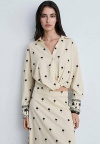 Chemise beige à boutonnières nouées avec broderie de palmiers noirs, associée à une jupe assortie présentant des motifs similaires et des poignets détaillés.